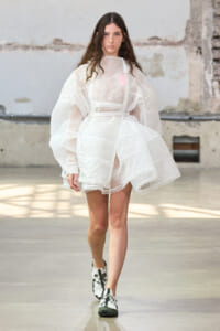 Model walking a white lace dress with oversized sleeves in an industrial setting, posing as a fashion show look.