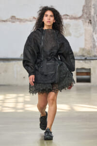 Model walking a runway in an all-black textured outfit with a belted waist and scalloped lace hem against an industrial backdrop.