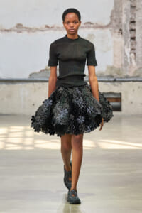 Female model walking a fashion show runway in a black fitted top and a voluminous black floral skirt.