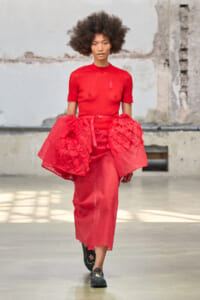Model walks the runway in a bold red ensemble featuring a fitted top and voluminous ruffled skirt in a textured red fabric, industrial backdrop behind.