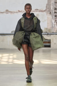 Model walking the runway in a black sheer hoodie and matching shorts, layered with olive-green cape panels and a green backpack with floral appliqués tied at the waist.