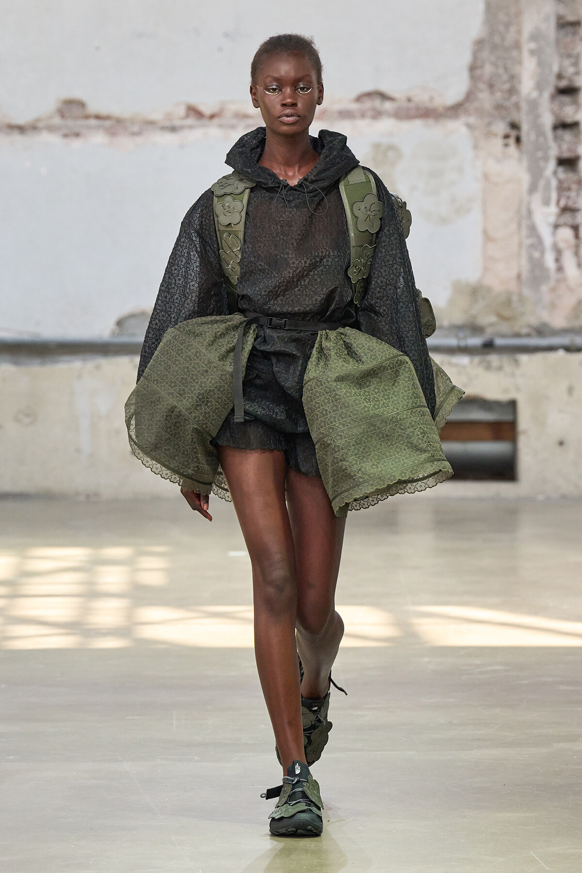 Model walking the runway in a black sheer hoodie and matching shorts, layered with olive-green cape panels and a green backpack with floral appliqués tied at the waist.