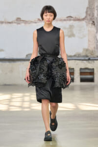 Model walking a runway in an all-black outfit: sleeveless top with a voluminous skirt featuring black floral appliqués and a belt.