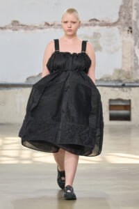 Model with platinum buzz cut wearing a black textured dress with wide straps, walking a fashion show on an industrial-style runway.