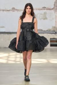 Model walking a runway in a black structured dress with thick straps and buckles, featuring a voluminous skirt against an industrial backdrop.