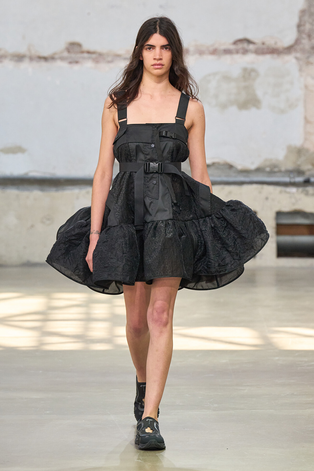 Model walking a runway in a black structured dress with thick straps and buckles, featuring a voluminous skirt against an industrial backdrop.