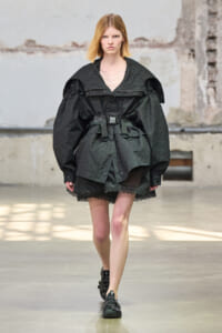 Female model walking the runway in a loose black utilitarian jacket with a belt over a black lace-skirt combo.