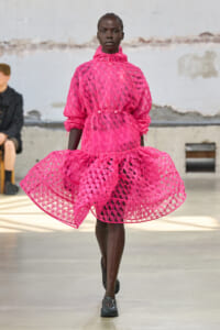 Model walking a fashion runway in a bright pink, lattice-cut dress with a high collar and voluminous skirt.