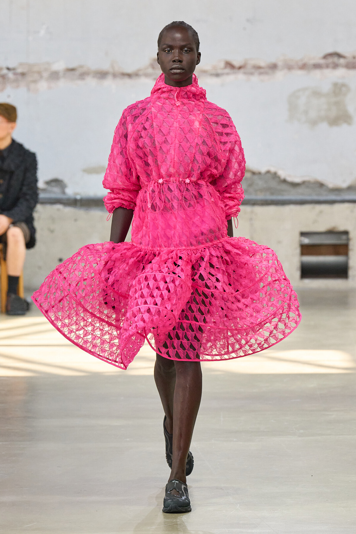 Model walking a fashion runway in a bright pink, lattice-cut dress with a high collar and voluminous skirt.