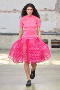 Woman walking toward camera in a pink knit top and bright pink lattice skirt, industrial backdrop behind her.