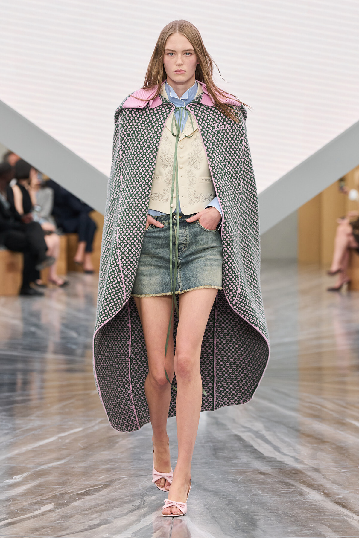 Model walking a fashion runway wearing a long patterned cape over a cream embroidered vest and denim shorts, pink accents visible at collar.