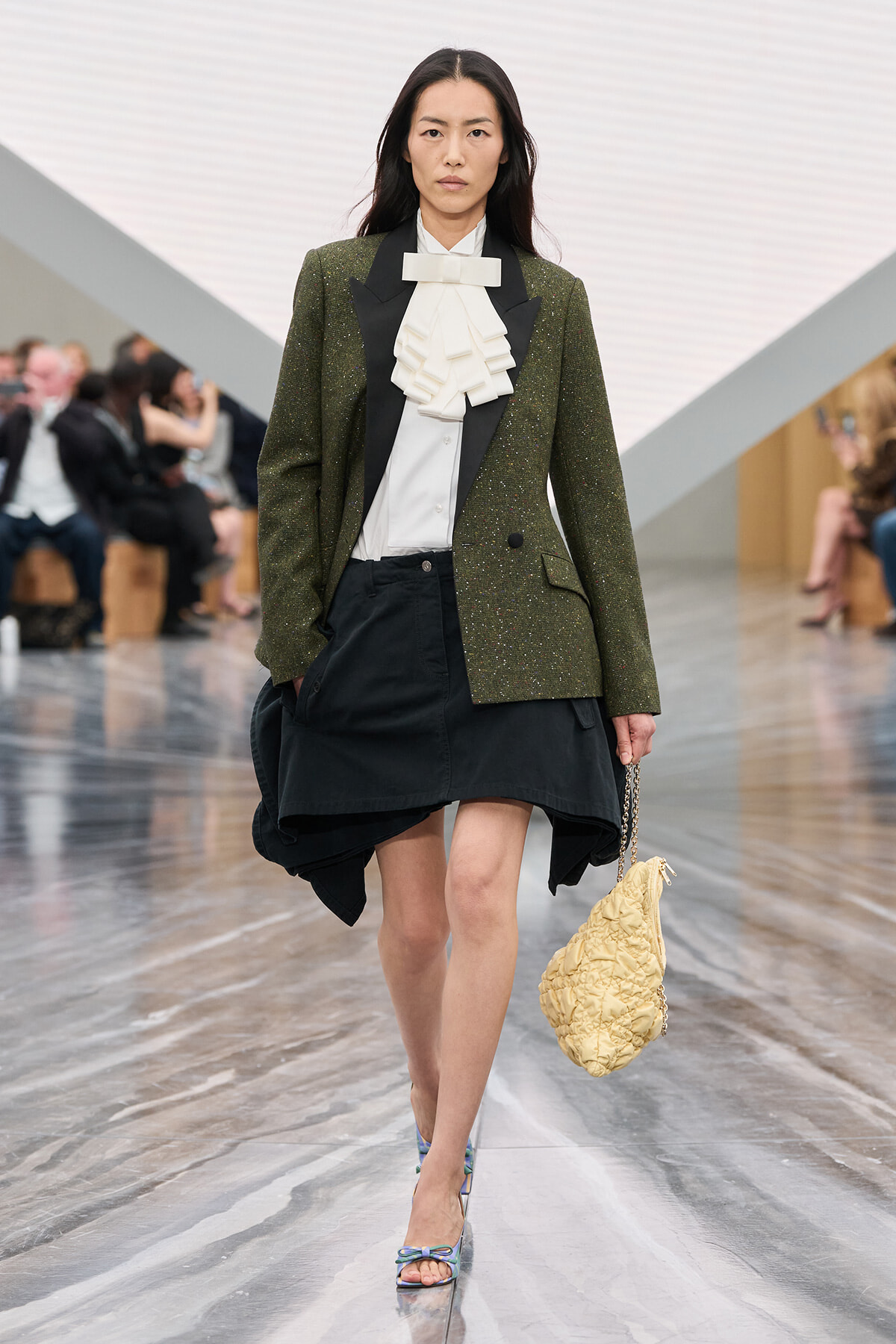 Model walks a runway wearing an olive glitter blazer over a white ruffled blouse and a black asymmetrical skirt, carrying a cream quilted bag with a chain strap.