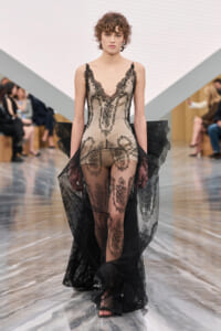Model on a runway wearing a sheer nude-toned lace bodysuit with black lace trim and a long black lace cape.