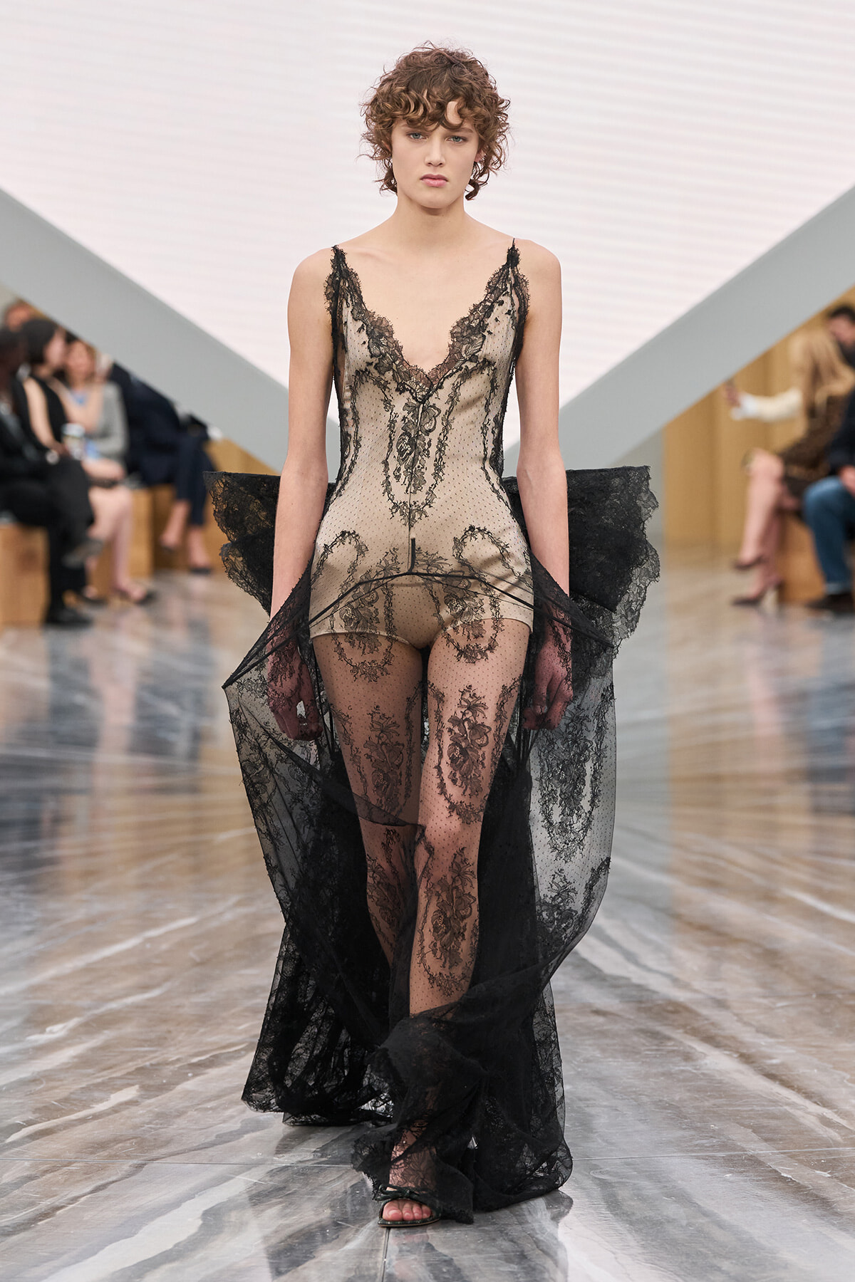 Model on a runway wearing a sheer nude-toned lace bodysuit with black lace trim and a long black lace cape.