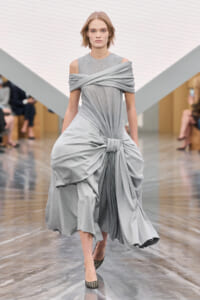 Model walks a fashion runway in a light gray, off-shoulder draped gown with a knotted waist.”] ,