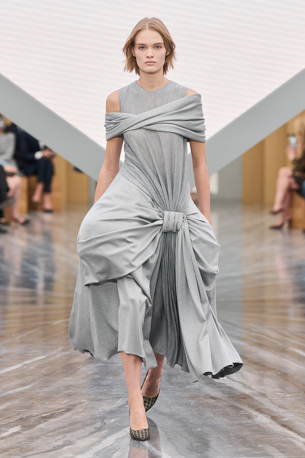 Model walks a fashion runway in a light gray, off-shoulder draped gown with a knotted waist.”] ,
