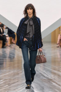 Model walking runway in a black textured coat with blue stripes, gray bow-tied scarf, dark jeans, and black loafers, carrying a brown quilted handbag.