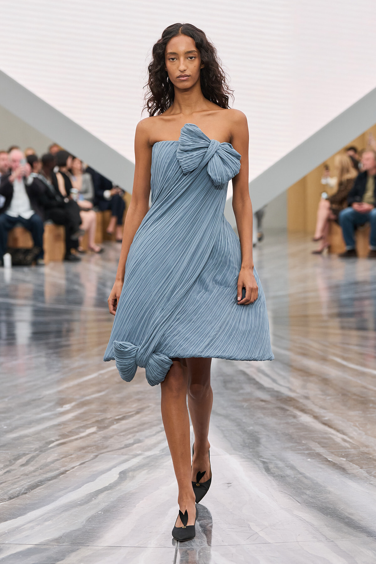 Model walks a runway in a light blue pleated strapless dress with oversized bows at the chest and hem, paired with black flats; audience seated along the sides.