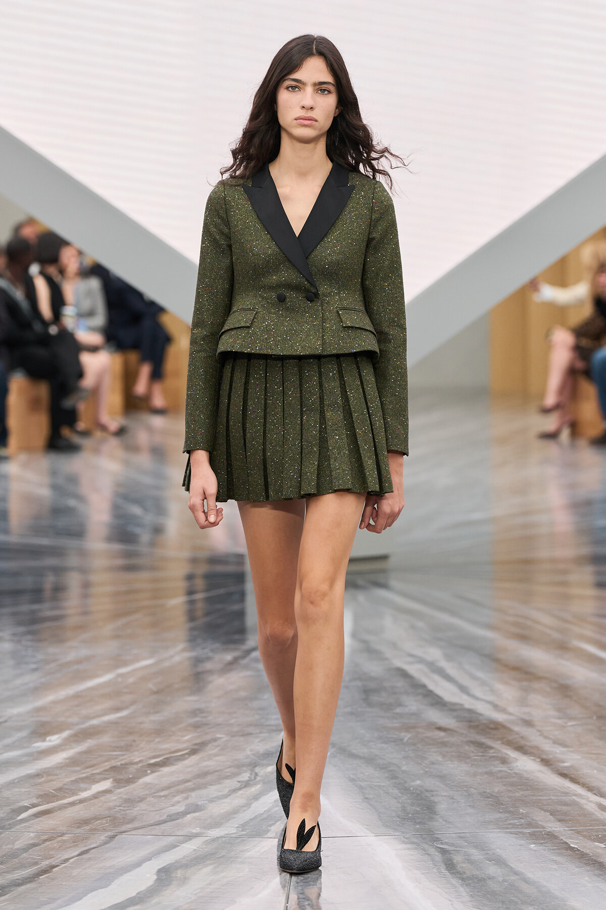 Model walking a runway in a sparkly olive-green blazer with black lapels and matching pleated mini skirt, wearing black heels on a glossy catwalk.