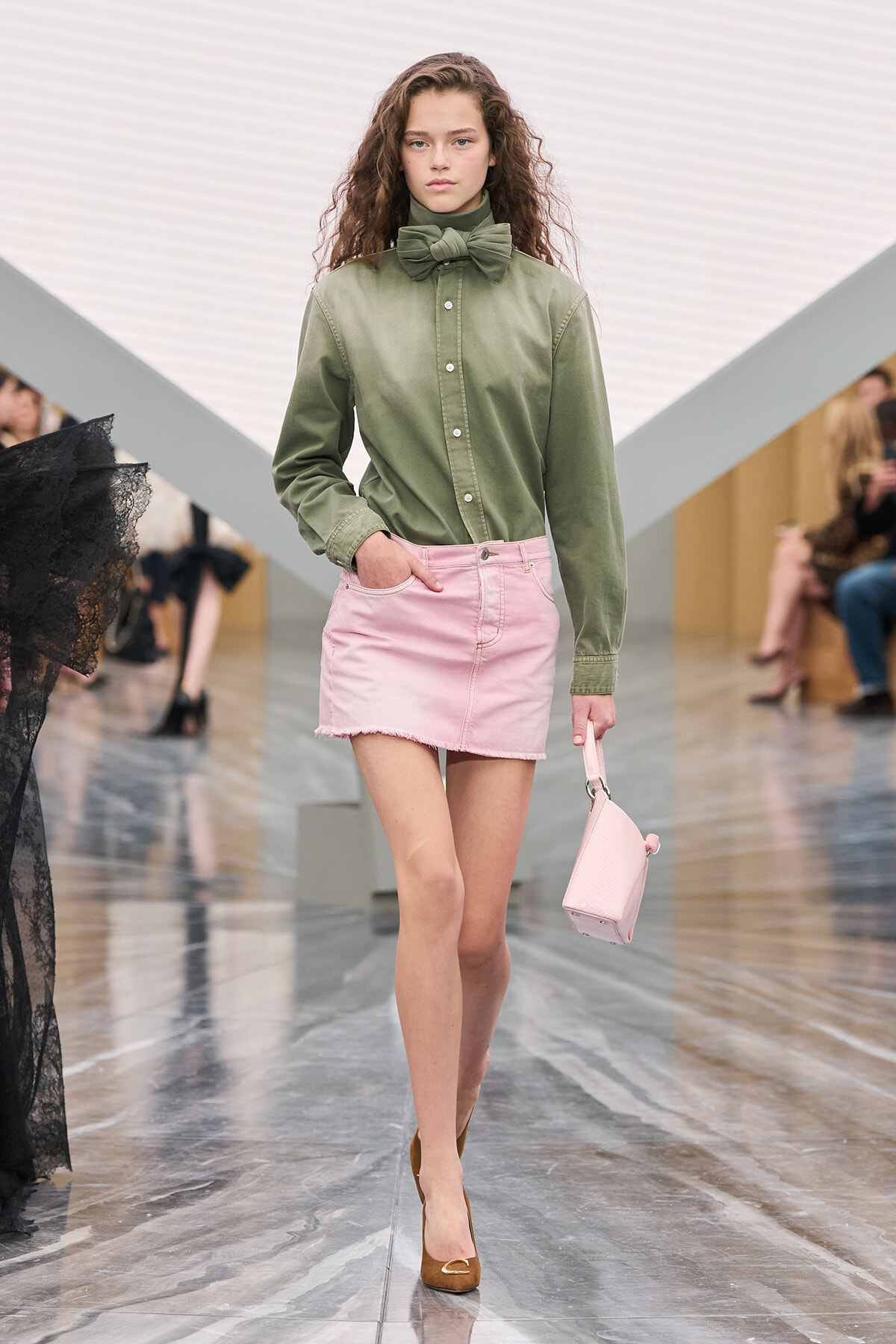 Model on a runway wearing an olive green button-down shirt with a large bow, a light pink frayed-hem mini skirt, brown heels, and a small pink handbag.