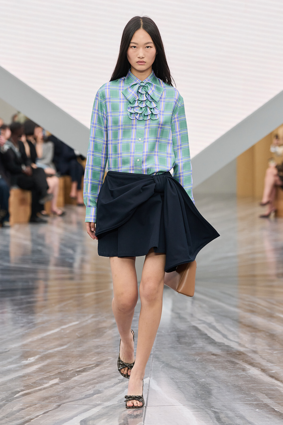 Model walking a runway in a pastel plaid blouse with a ruffled bow and a navy wrap skirt, carrying a tan clutch.