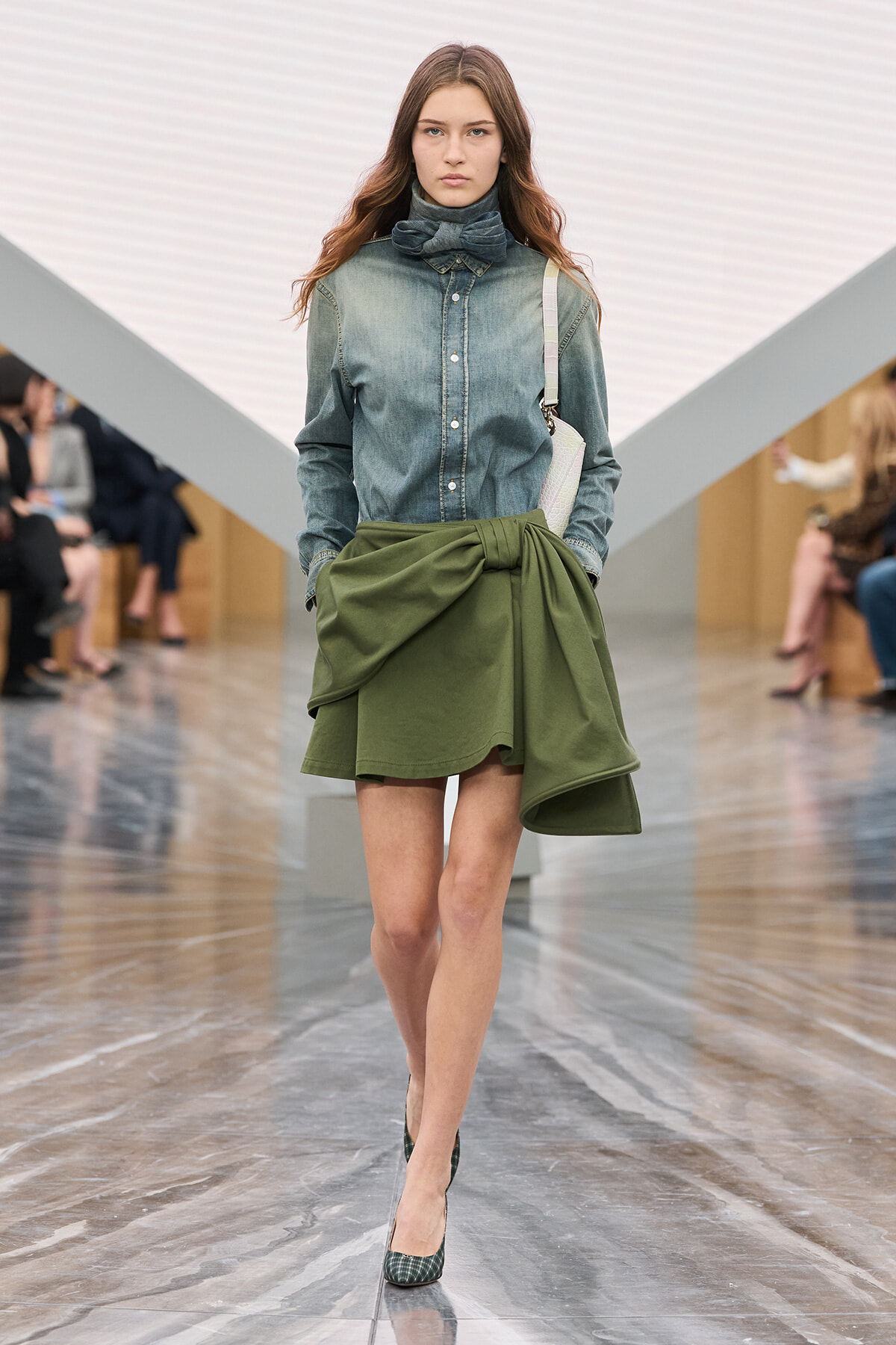 Model walking a runway in a blue denim button-up shirt, an olive-green draped wrap skirt, and green patterned heels with a light bag over the shoulder.