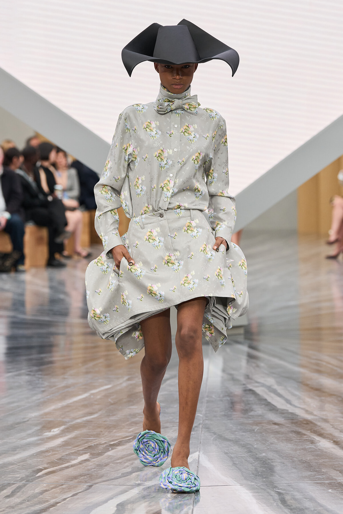 Model on a runway wearing a light gray floral shirt dress with a matching bow, an oversized black sculptural hat, and wraparound blue-green flower-shaped slippers.
