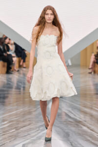 Model walking a runway in a white strapless dress adorned with circular lace appliqués on an icy-toned set.