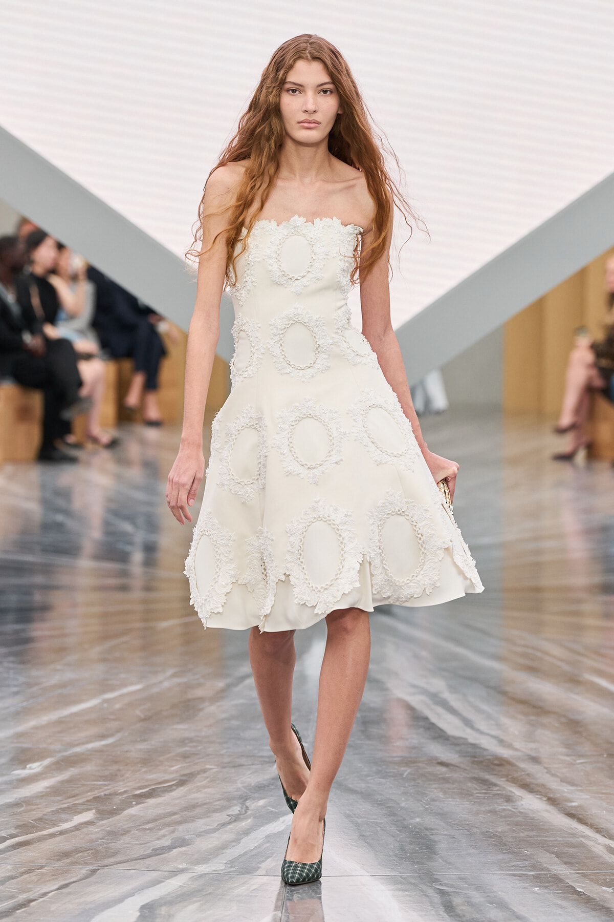 Model walking a runway in a white strapless dress adorned with circular lace appliqués on an icy-toned set.