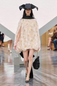 Model walking the runway in a beige floral cape dress with a dramatic oversized black hat and a black quilted bag at her side.