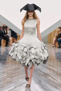Model on a runway wearing a silver sleeveless gown with a voluminous skirt made of stacked rectangular panels and a large black sculptural hat.