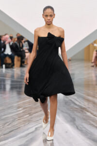 Model on a runway wearing a black, pleated strapless dress with a large bow at the chest and a bow at the hem, paired with white heels.
