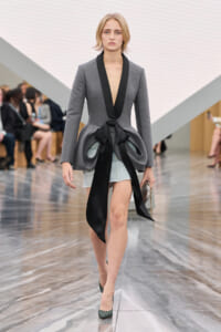 Runway model wearing a gray tailored blazer with black satin lapels and a large black bow over a denim mini skirt, holding a clutch.