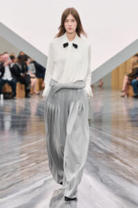 Model walking runway in a white blouse with black bow and voluminous grey pleated pants, hands in pockets, audience in background