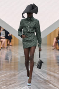 Model walks the runway in a green checkered mini dress with a high-neck bow, paired with an oversized sculptural black hat and a small black handbag.
