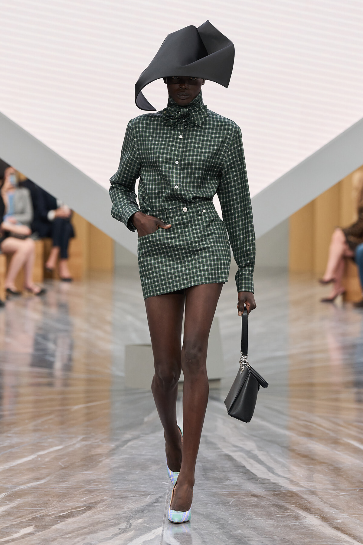 Model walks the runway in a green checkered mini dress with a high-neck bow, paired with an oversized sculptural black hat and a small black handbag.