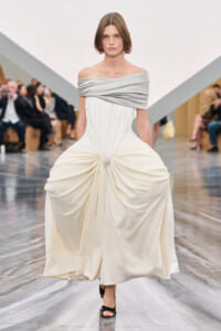 Model walking a runway in an off-shoulder ivory gown with a draped gray neckline and center knot, black heels, audience in the background.