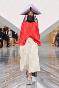 Model on a runway wearing a large lavender sculptural hat, red pleated top, and voluminous cream pants with a black lace veil.
