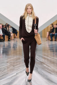 Model walks a fashion runway wearing a dark brown matching set and cream ruffled blouse, carrying a tan shoulder bag and black heels with gold accents