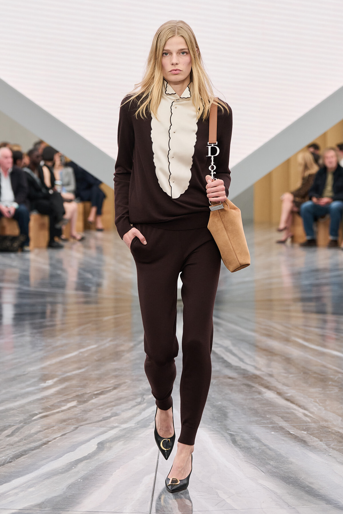 Model walks a fashion runway wearing a dark brown matching set and cream ruffled blouse, carrying a tan shoulder bag and black heels with gold accents