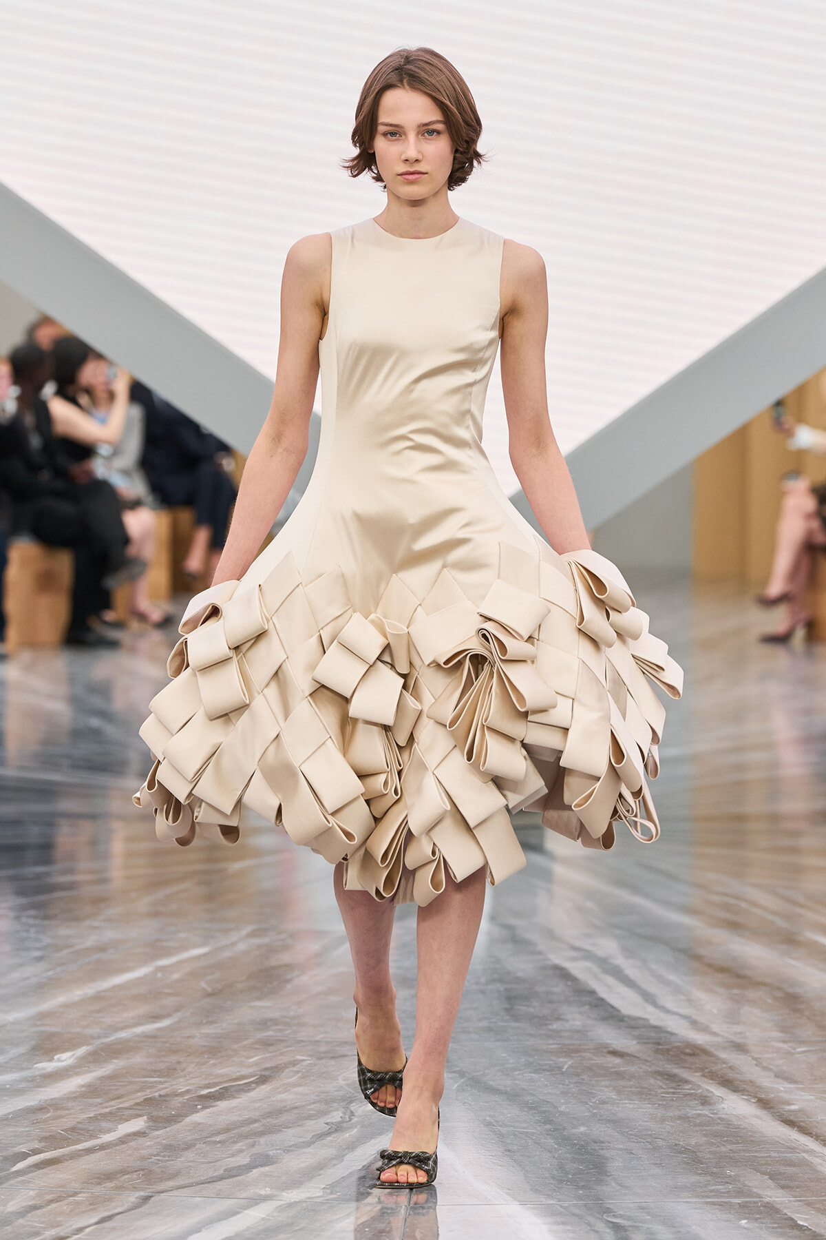 Model walking a runway in a sleeveless cream dress with a voluminous geometric ruffle skirt formed from square folds.