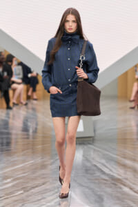 Runway model in a navy blue plaid shirt dress with a large bow, carrying a brown suede tote with a Dior strap.