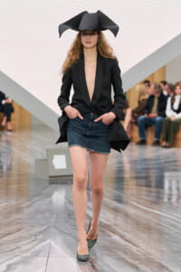 Model walks a runway in a black blazer with deep plunging neckline, a denim mini skirt, green checkered heels, and a large avant-garde black hat; audience seated in background