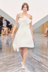 Model walking a white strapless A-line dress with a large bow at the bust and a bow at the hem on a fashion show runway, audience blurred in background, overall pale setting.