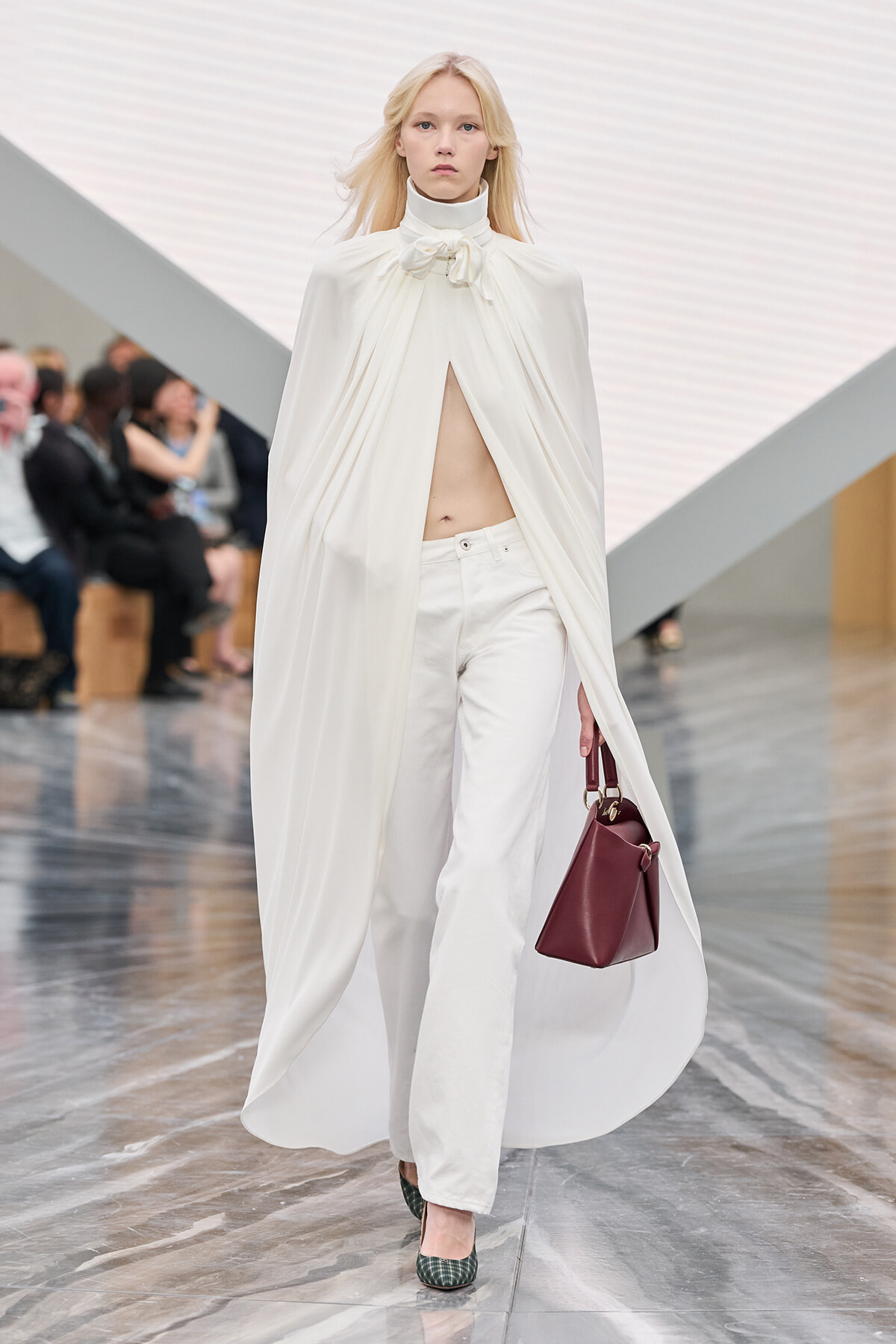 Female model on a runway in an all-white ensemble: flowing cape top, wide white pants, and a burgundy handbag.