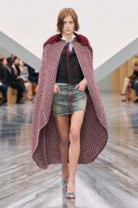 Model walks the runway in a long burgundy patterned cape over a black vest and light-blue blouse, paired with frayed denim shorts and pastel sandals, demonstrating a layered fashion look.