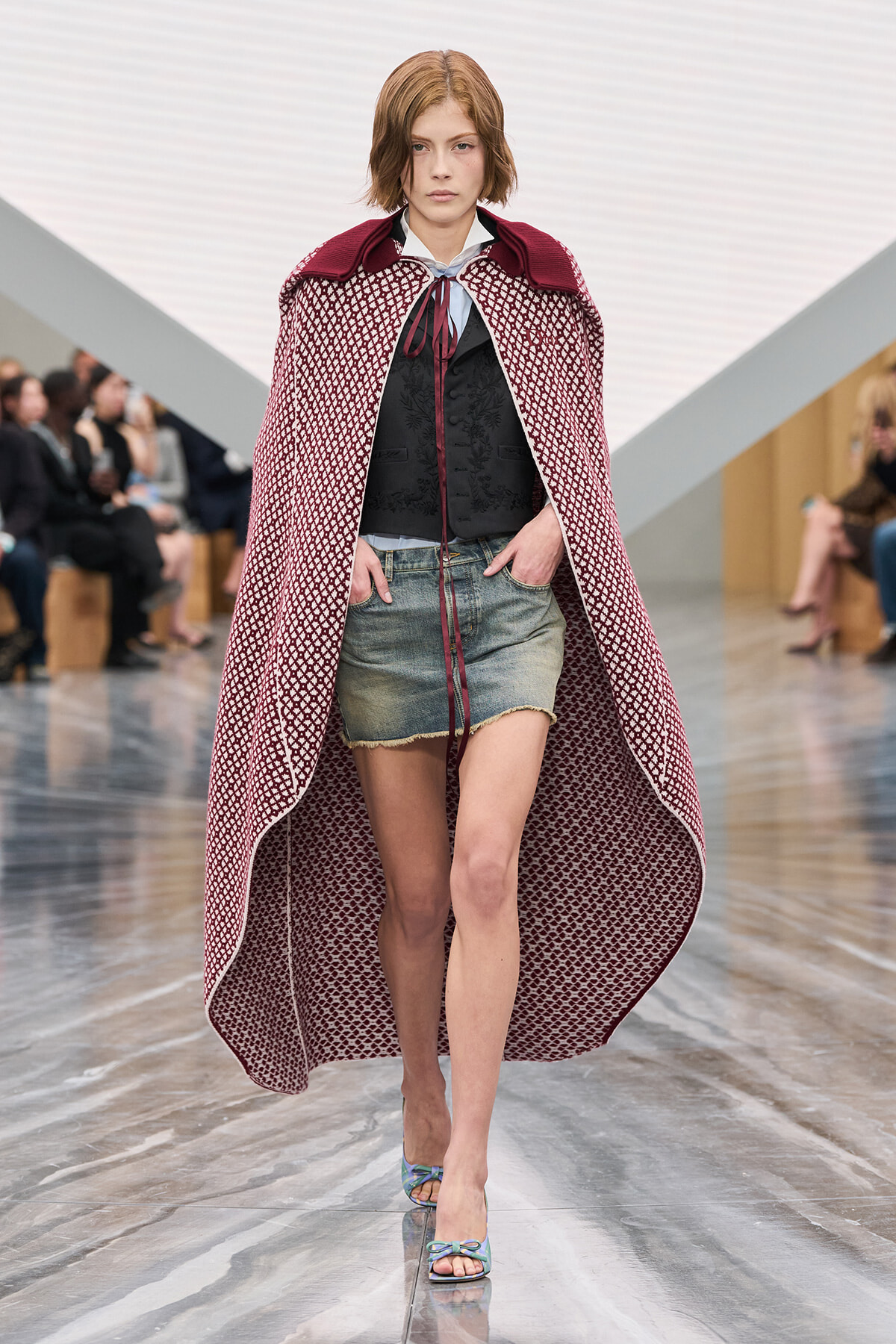 Model walks the runway in a long burgundy patterned cape over a black vest and light-blue blouse, paired with frayed denim shorts and pastel sandals, demonstrating a layered fashion look.