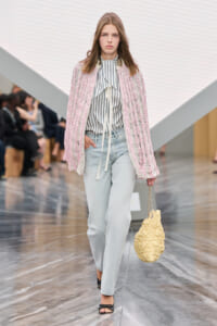 Model walks a fashion runway in a pink textured cardigan over a striped blouse, light jeans and black sandals, carrying a yellow quilted bag with a chain.