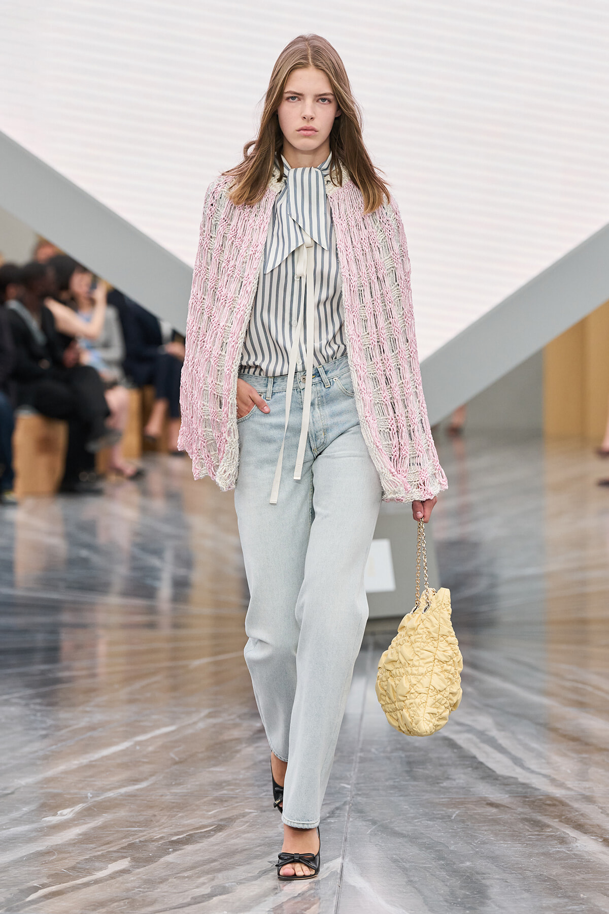 Model walks a fashion runway in a pink textured cardigan over a striped blouse, light jeans and black sandals, carrying a yellow quilted bag with a chain.