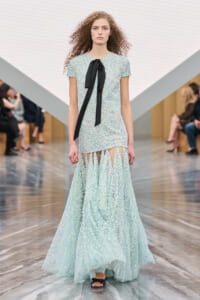 Model walks a runway wearing a pale blue, textured gown with a black silk bow at the neckline